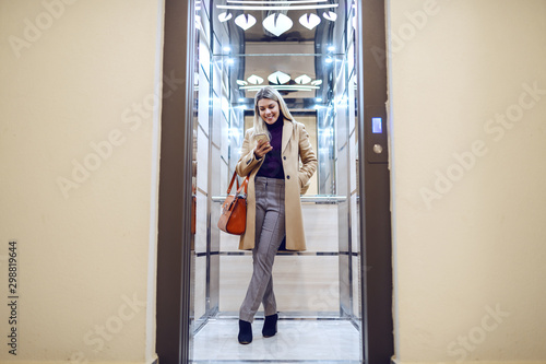 Full length of cheerful caucasian blonde woman in coat standing in elevator and using smart phone. photo