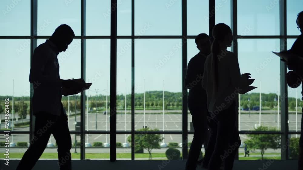 Silhouette of businessman standing by window in office hall, drinking coffee at break and thinking. Office workers walking by in slow motion Silhouette of businessman standing by window in office hall, drinking coffee at break and thinking. Office workers walking by in slow motion