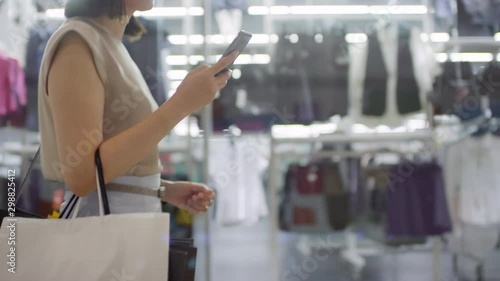 Tracking shot of unrecognizable woman with short hair walking through shopping mall past glass window displays, using smartphone and carrying many paper bags with purchases photo