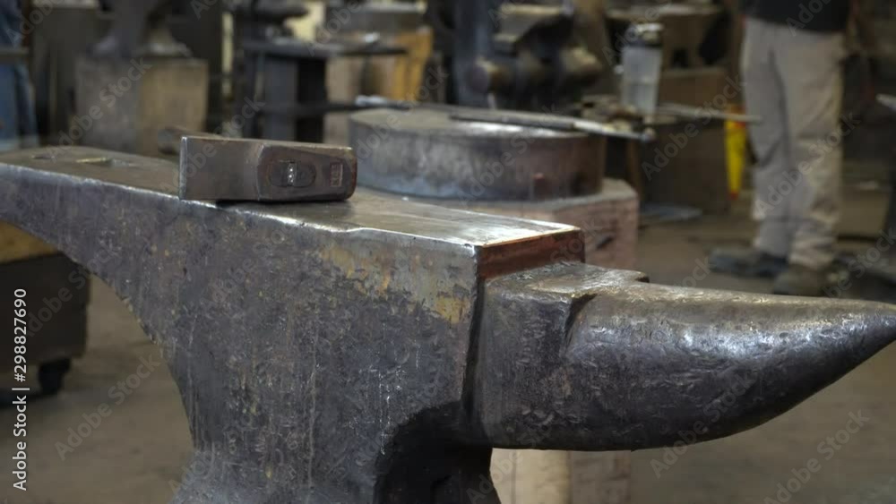 We can see a hammer on an anvil in a blacksmith factory. The blacksmiths are moving a lot of behind the anvil. We can see a hammer on an anvil in a blacksmith factory. The blacksmiths are moving a lot of behind the anvil.