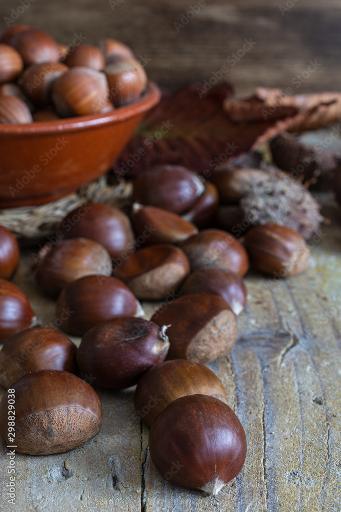 Close-up of chestnuts with leaves and ceramic bowl with unfocused hazelnuts, on weathered wooden background in vertical Close-up of chestnuts with leaves and ceramic bowl with unfocused hazelnuts, on weathered wooden background in vertical