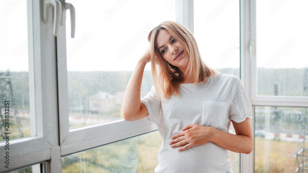 Portrait of a pregnant happy woman standing at panoramic window on a fresh rainy autumn day, smiling at her thoughts. Portrait of a pregnant happy woman standing at panoramic window on a fresh rainy autumn day, smiling at her thoughts.