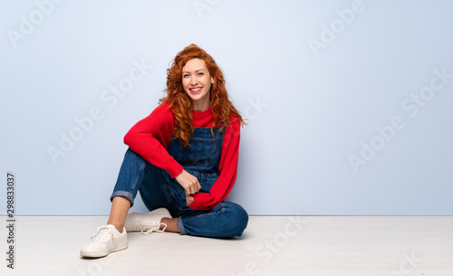 Redhead woman with overalls sitting on the floor posing with arms at hip and smiling