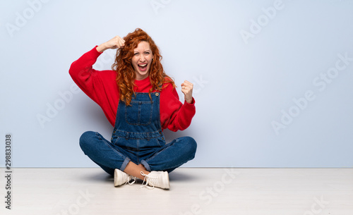 Redhead woman with overalls sitting on the floor celebrating a victory