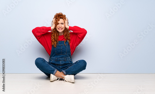 Redhead woman with overalls sitting on the floor frustrated and takes hands on head