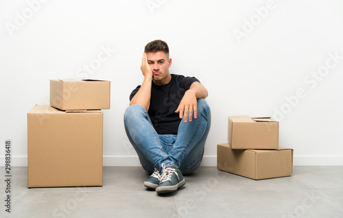 Handsome young man moving in new home among boxes unhappy and frustrated