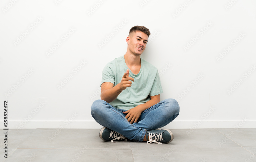 Young handsome man sitting on the floor points finger at you