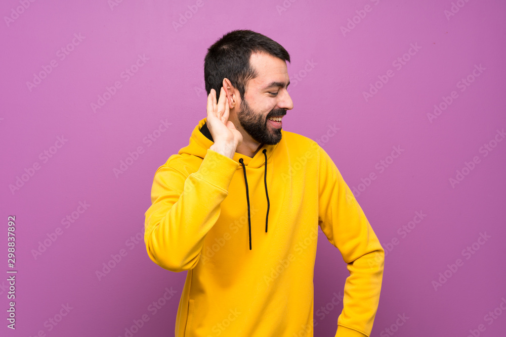 Fototapeta premium Handsome man with yellow sweatshirt listening to something by putting hand on the ear