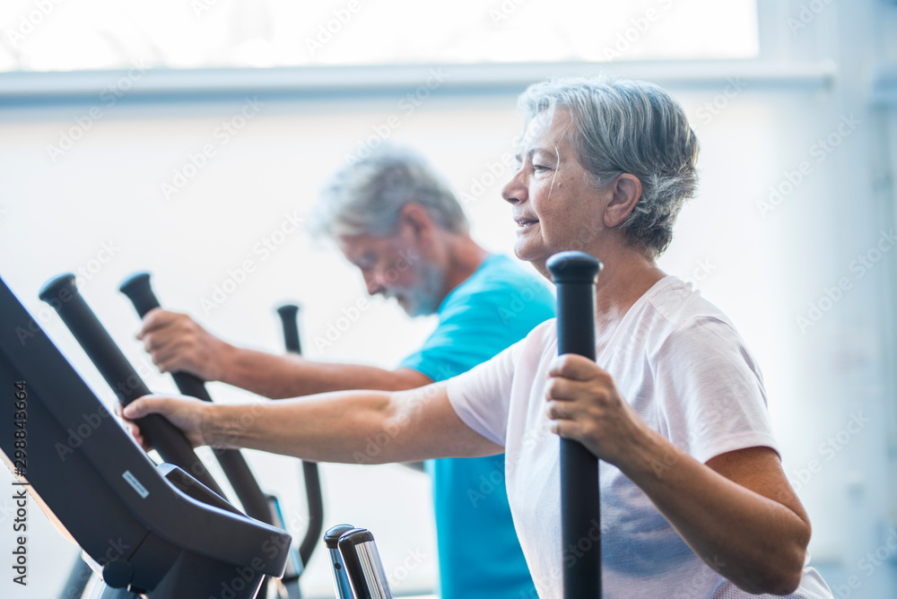 woman using a precor at the gym - active senior with his man at the background - couple of pensioners doing exercises together woman using a precor at the gym - active senior with his man at the background - couple of pensioners doing exercises together