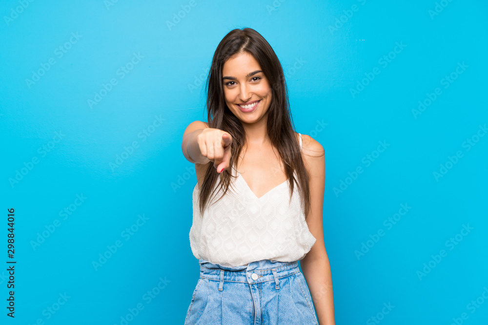 Fototapeta premium Young woman over isolated blue background points finger at you while smiling