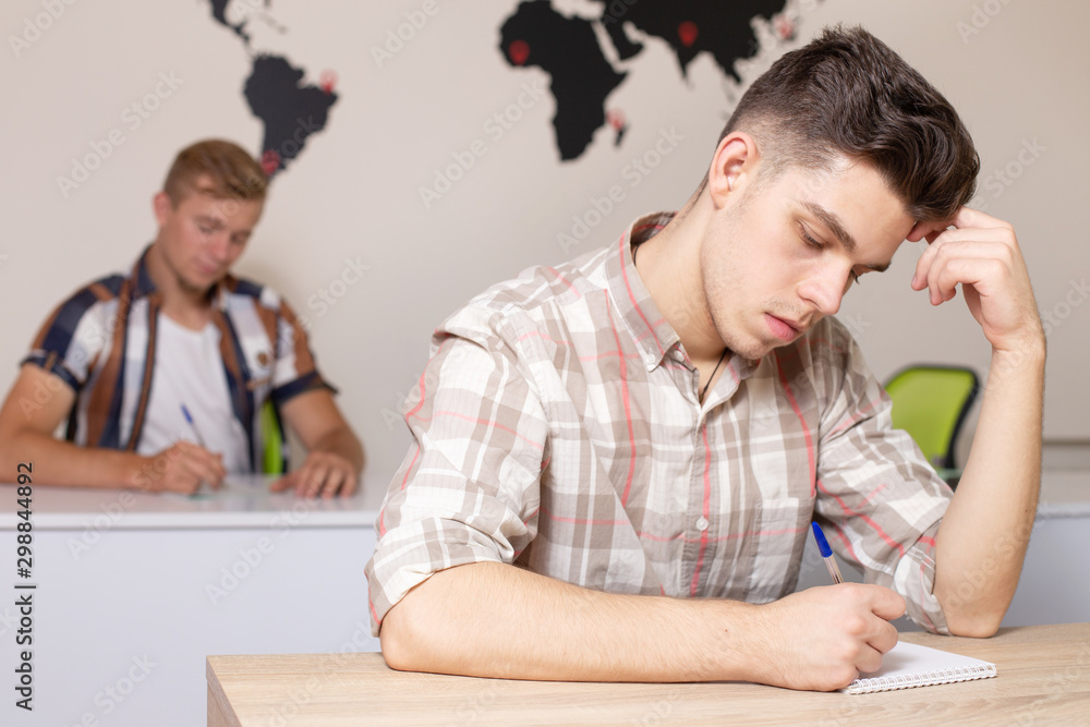Students in classroom with world map on the wall on background. Young guy writes an abstract or does a test. Education, university, learning, coworking concept Students in classroom with world map on the wall on background. Young guy writes an abstract or does a test. Education, university, learning, coworking concept