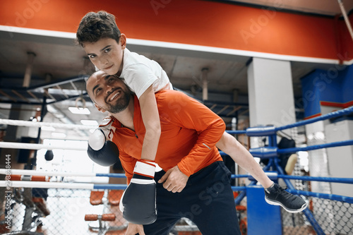 Fototapeta Naklejka Na Ścianę i Meble -  cheerful young bearded man giving piggyback ride to smiling kid in boxing gloves at gym, free time, spare time, lifestyle