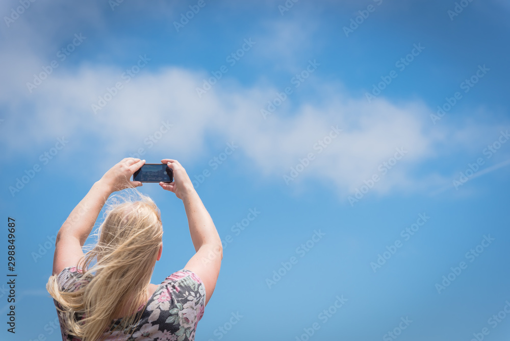 Young Caucasian lady taking photo using her smart phone against blue sky Young Caucasian lady taking photo using her smart phone against blue sky