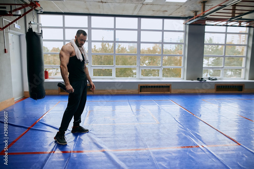 tired muscular bodybuilder with water bottle and towel on his shoulder looking down having a rest. copy space. full length photo. tiredness, exhaustion, punching bag in the background of the photo