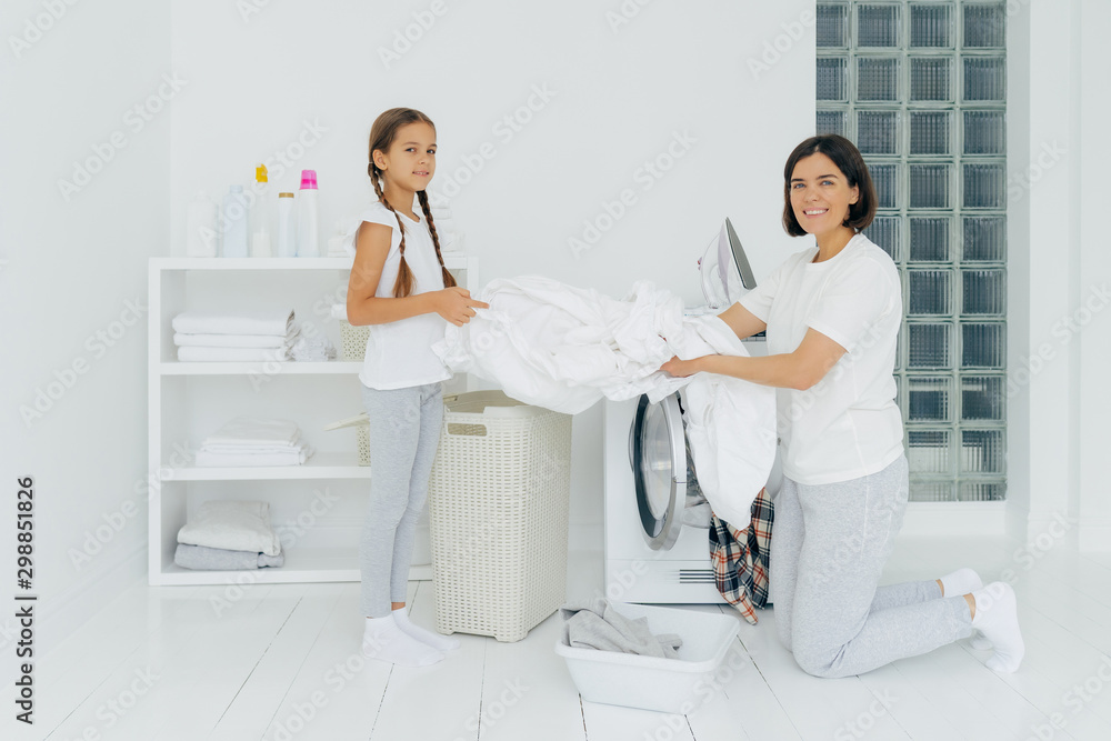 Obraz premium Glad housewife does washing with little adorable helper. Mother and daughter wash clothes in laundry room, load linen in washer. Woman stands on knees near washing machine. Housework concept