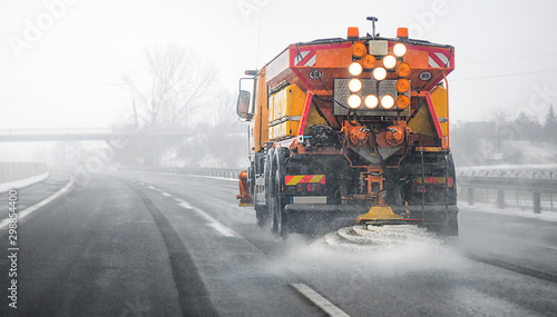 Snow plow salting street in winter time. Orange truck deicing. Maintenance winter vehicle back side.