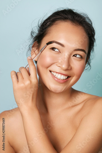 Makeup. Woman brushing brows with eyebrow brush closeup. Portrait of happy smiling asian girl model with beautiful face shaping eyebrows with cosmetic brush at studio