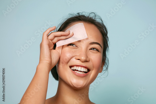 Face skin care. Smiling woman using facial oil blotting paper portrait. Closeup of beautiful happy asian girl model with natural makeup using oil absorbing sheets, beauty product at studio