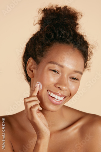 Face skin care. Woman applying cosmetic cream on clean hydrated skin portrait. Beautiful happy smiling african american girl model with natural makeup applying facial moisturizer, beauty product