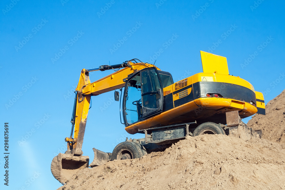 A yellow black excavator standing on a large pile of ground against a blue sky. Digger with a bucket. Road work on an intercity highway