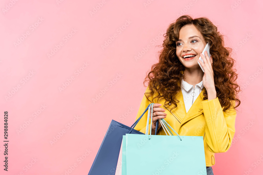 Fototapeta premium beautiful curly girl holding shopping bags while talking on smartphone, isolated on pink
