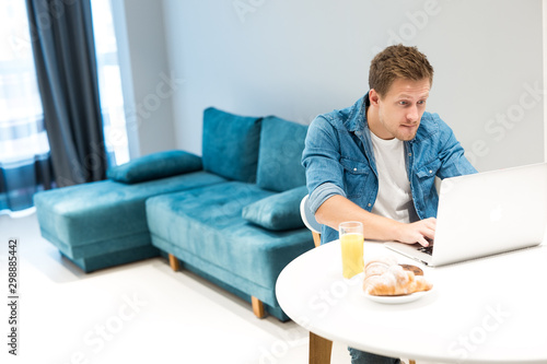 young handsome man working in his laptop from home while having croissant and fresh orange juice for lunch looking excited