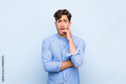 Fotografija young handsome man open-mouthed in shock and disbelief, with hand on cheek and a