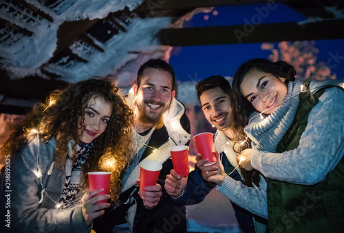 Group of young friends outdoors in snow in winter at night, holding drinks.