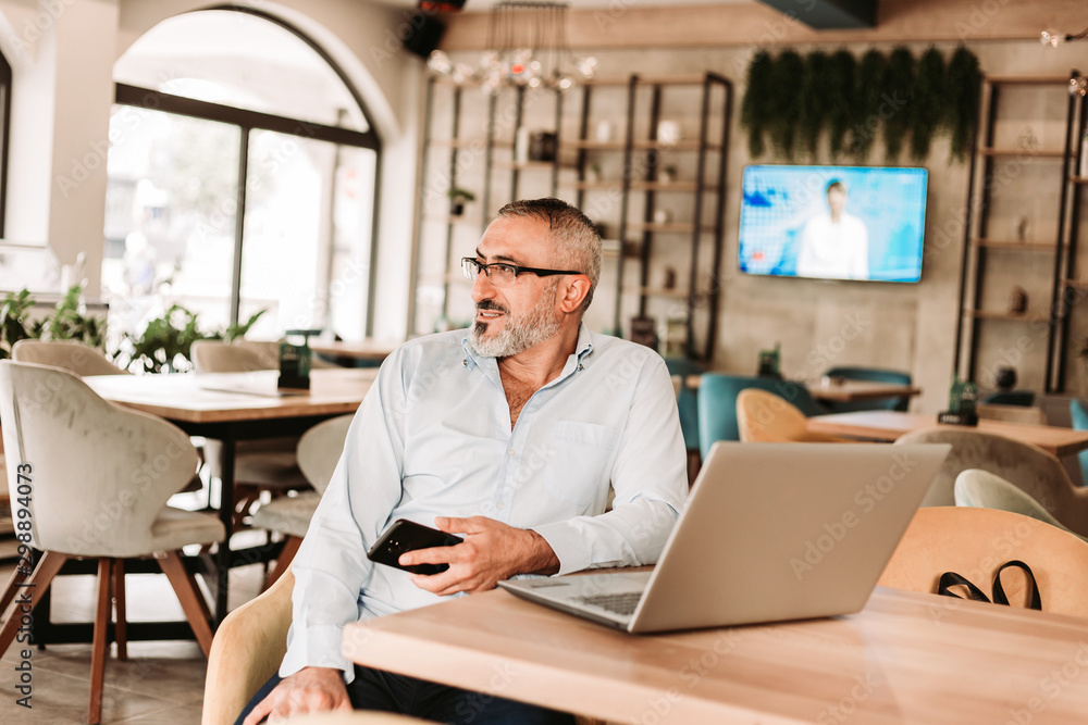 Middle aged man working on laptop in cafe, holding phone in his hand Middle aged man working on laptop in cafe, holding phone in his hand