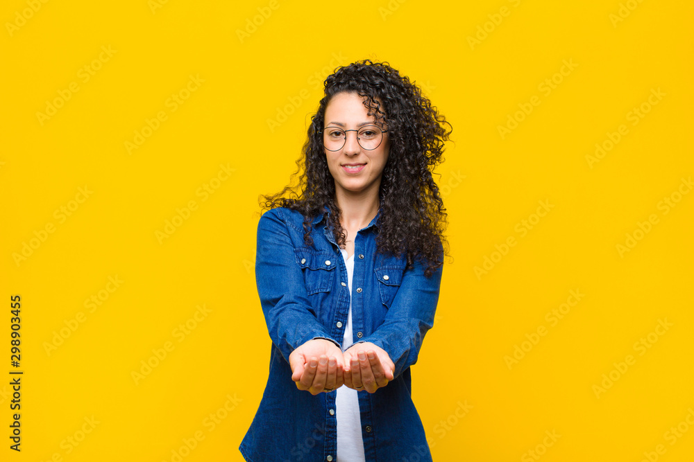 young pretty woman smiling happily with friendly, confident, positive look, offering and showing an object or concept against orange wall - obrazy, fototapety, plakaty 