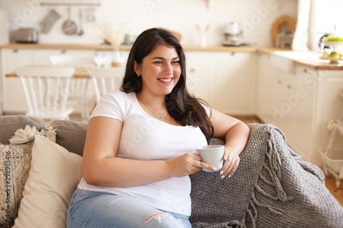 Portrait of cheerful charismatic young overweight female with big breast and long black hair posing in stylish kitchen interior, having coffee on comfortable couch and laughing at funny joke photo