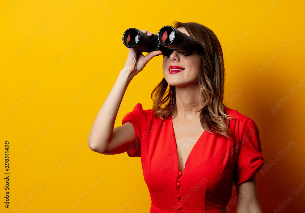 Young woman in red dress with binocular on yellow background Young woman in red dress with binocular on yellow background