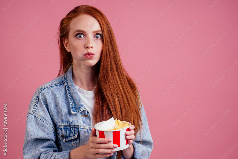 cute red long foxy haired ginger woman holding packaging cardboard box bucket with french fries and eating chicken crunchy nuggets in pink studio cute red long foxy haired ginger woman holding packaging cardboard box bucket with french fries and eating chicken crunchy nuggets in pink studio