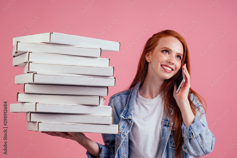 pretty long ginger red-haired woman holding a lot of boxes with fast food snack and smartphone isolated pink background studio shot. delivery for home party concept pretty long ginger red-haired woman holding a lot of boxes with fast food snack and smartphone isolated pink background studio shot. delivery for home party concept