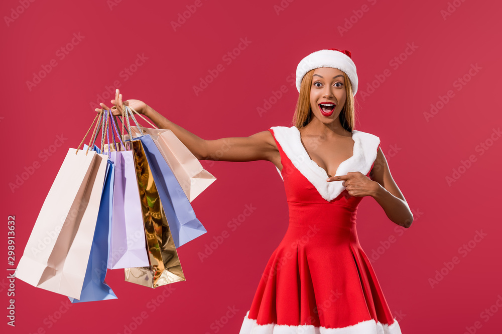 excited african american girl in santa hat and christmas dress pointing with finger at shopping bags isolated on red