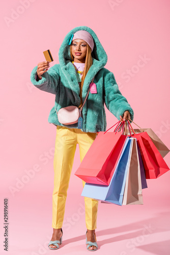 stylish african american woman holding credit card and shopping bags on pink background, fashion doll concept