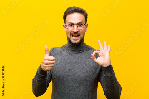 young handsome man feeling happy, amazed, satisfied and surprised, showing okay and thumbs up gestures, smiling against orange wall