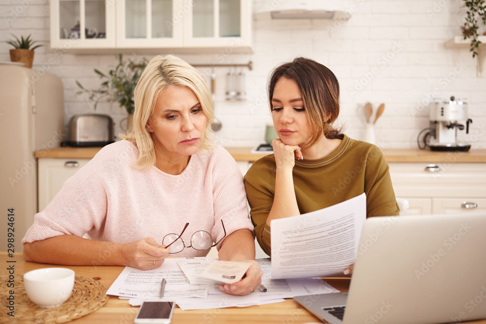 Picture of friendly caring young dark haired woman helping mother to fill in financial papers at home. Attractive blonde middle aged female is taught to pay utility bills online using laptop Picture of friendly caring young dark haired woman helping mother to fill in financial papers at home. Attractive blonde middle aged female is taught to pay utility bills online using laptop