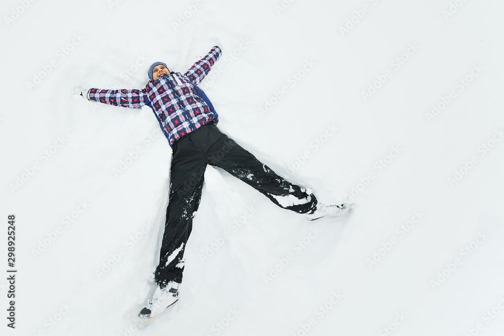 Fototapeta premium young man in a plaid outerwear or jacket lies on the snow and makes the figure of an angel. Winter fun