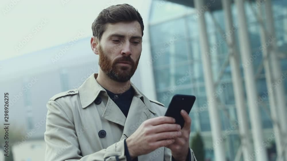 Portrait of Handsome Man is using his Phone after he smiles to the Camera. Attractive Businessman looks charmingly to the Camera. He dressed classical Suit. Smartphones. People. Men. Apps. Portrait of Handsome Man is using his Phone after he smiles to the Camera. Attractive Businessman looks charmingly to the Camera. He dressed classical Suit. Smartphones. People. Men. Apps.