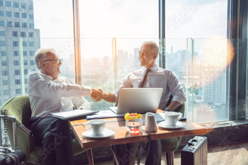 Two businessmen shaking hands together while sitting by windows.Mature businessman discuss information with a colleague in a modern business lounge high up in an office tower overlooking the city.