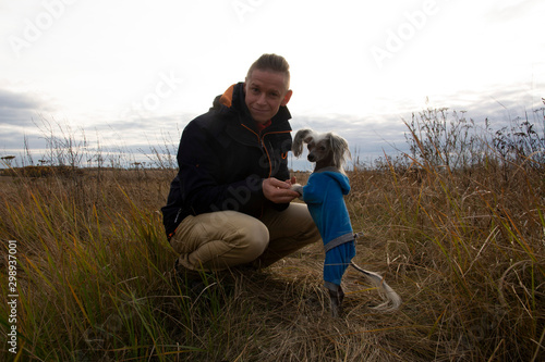A middle-aged man walks with a dog breed Chinese Hairless Crested Dog on nature in autumn afternoon.