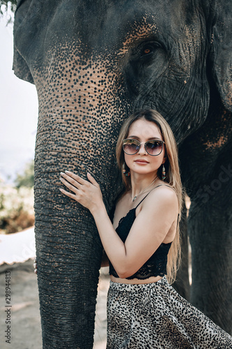 Pretty blond woman in stylish sunglasses and a dress with leopard print gently hugs elephant’s trunk; friendship concept.