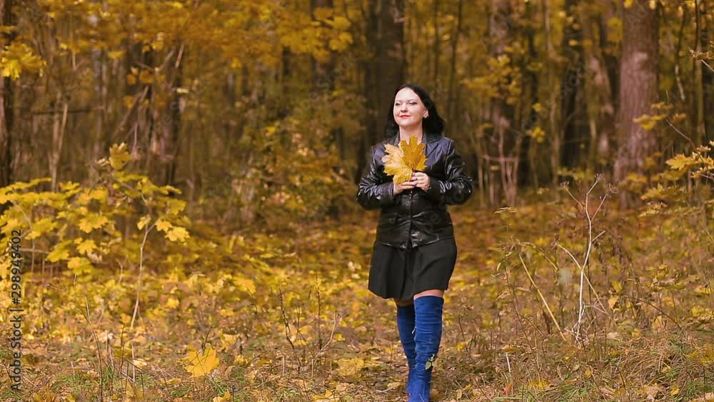 A young brunette woman in blue boots is walking in an autumn park with yellow leaves in her hands A young brunette woman in blue boots is walking in an autumn park with yellow leaves in her hands