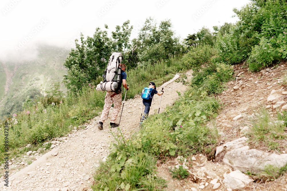 Obraz premium Dad and son hiking with heavy backpacks climb a mountain trail. Parenthood