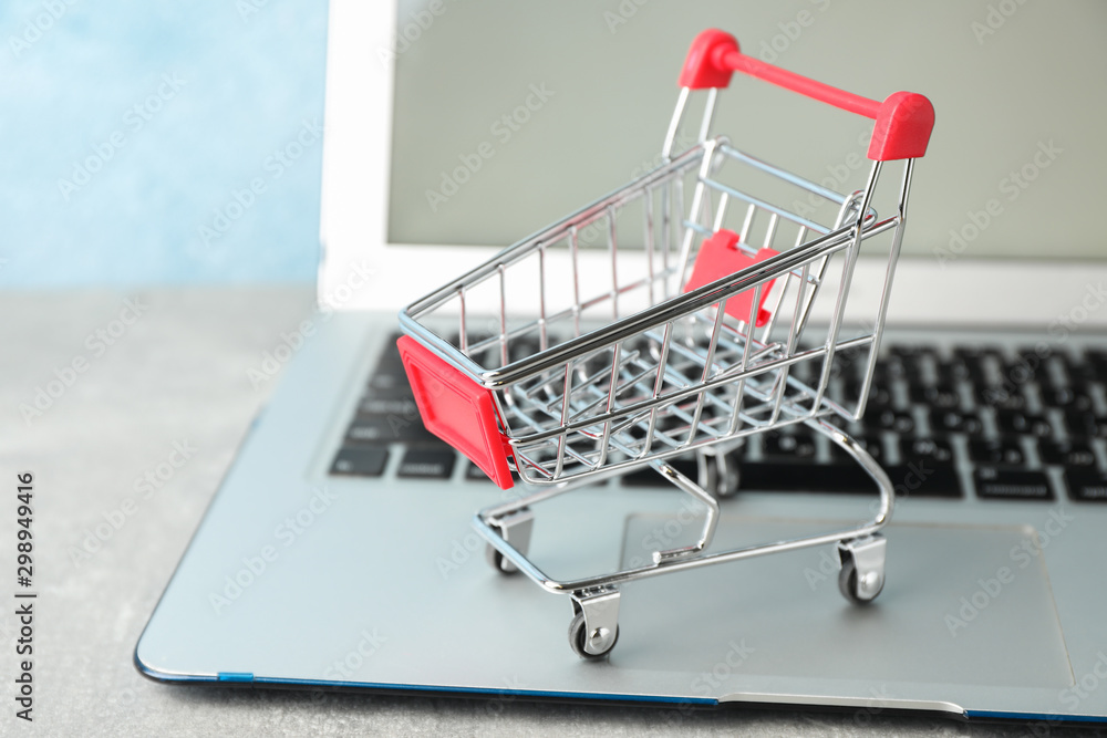 Small shopping cart and laptop on grey background, copy space - obrazy, fototapety, plakaty 