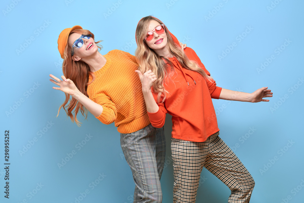 Two Lovable embracing fashionable woman sisters having fun jump in Trendy orange outfit. Studio shot of Carefree stylish friends laughing on blue background. Cheerful fashion girl, funny mood