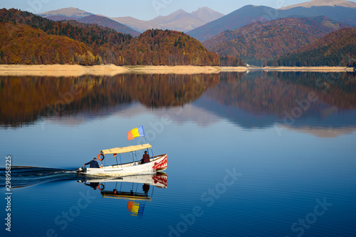 Sailing boat with water reflection on Vidraru Dam, Romania.  On a background of beautiful colorful autumn forest with red yellow and green trees in sunny day. 