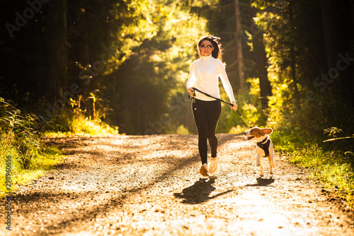 Girl is running with a dog (Beagle) on a leash in the autumn time, sunny day in forest. Copy space in nature