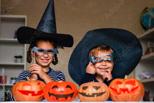 Happy brother and sister celebrate Halloween. Funny children in carnival costumes indoors at the table with pumpkins. Cheerful children play with pumpkins and masks on sticks